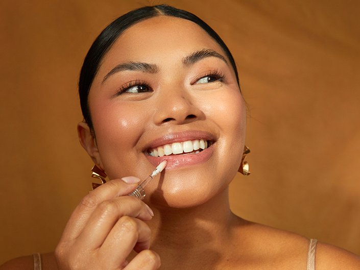 Person smiling with a natural makeup look and hair pulled back off of their face applies clear lip gloss to their lips