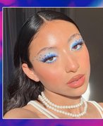 Person with black hair wearing blue eyeshadow with blue rhinestones, nude lipstick and a pearl necklace