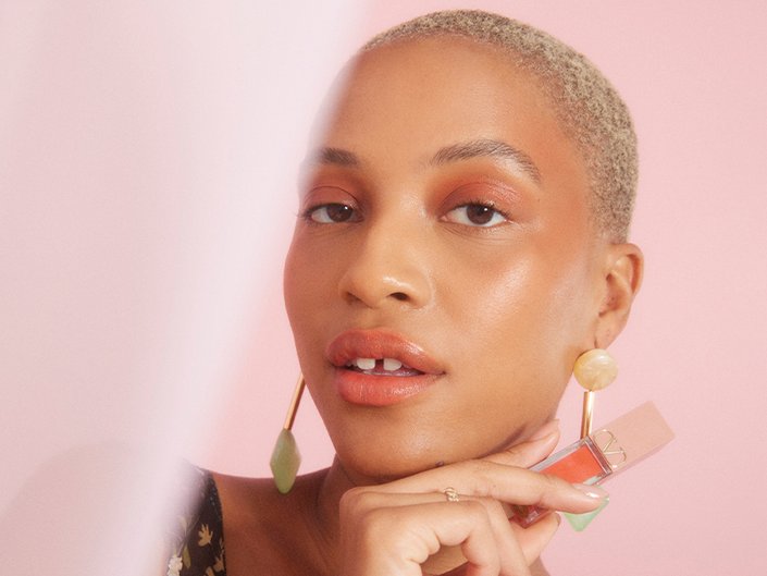 Picture of a model with bleached blonde buzzcut holding a lip product on a soft pink background