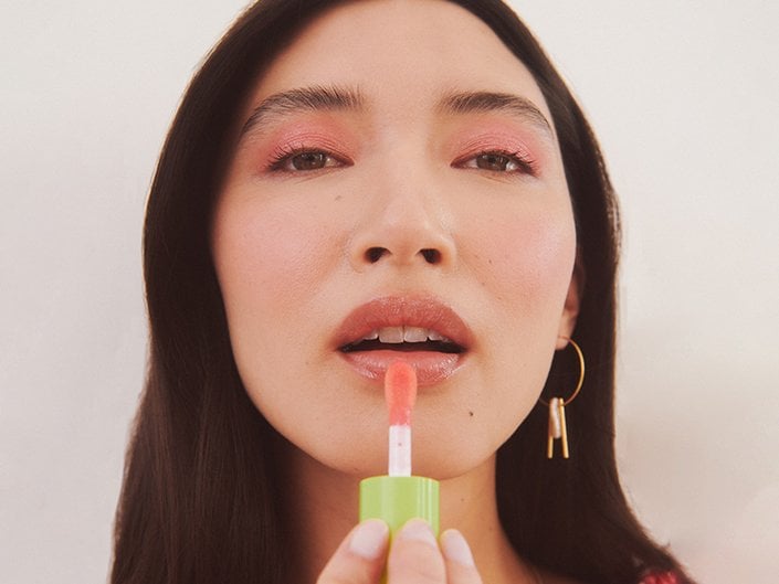 A model with dark hair and pastel pink eyeshadow applying a pale pink shade of the NYX Professional Makeup Fat Oil Lip Drip to their lips.  