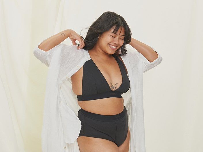 Person with long dark hair smiling and wearing a black bra and underwear with a white shirt